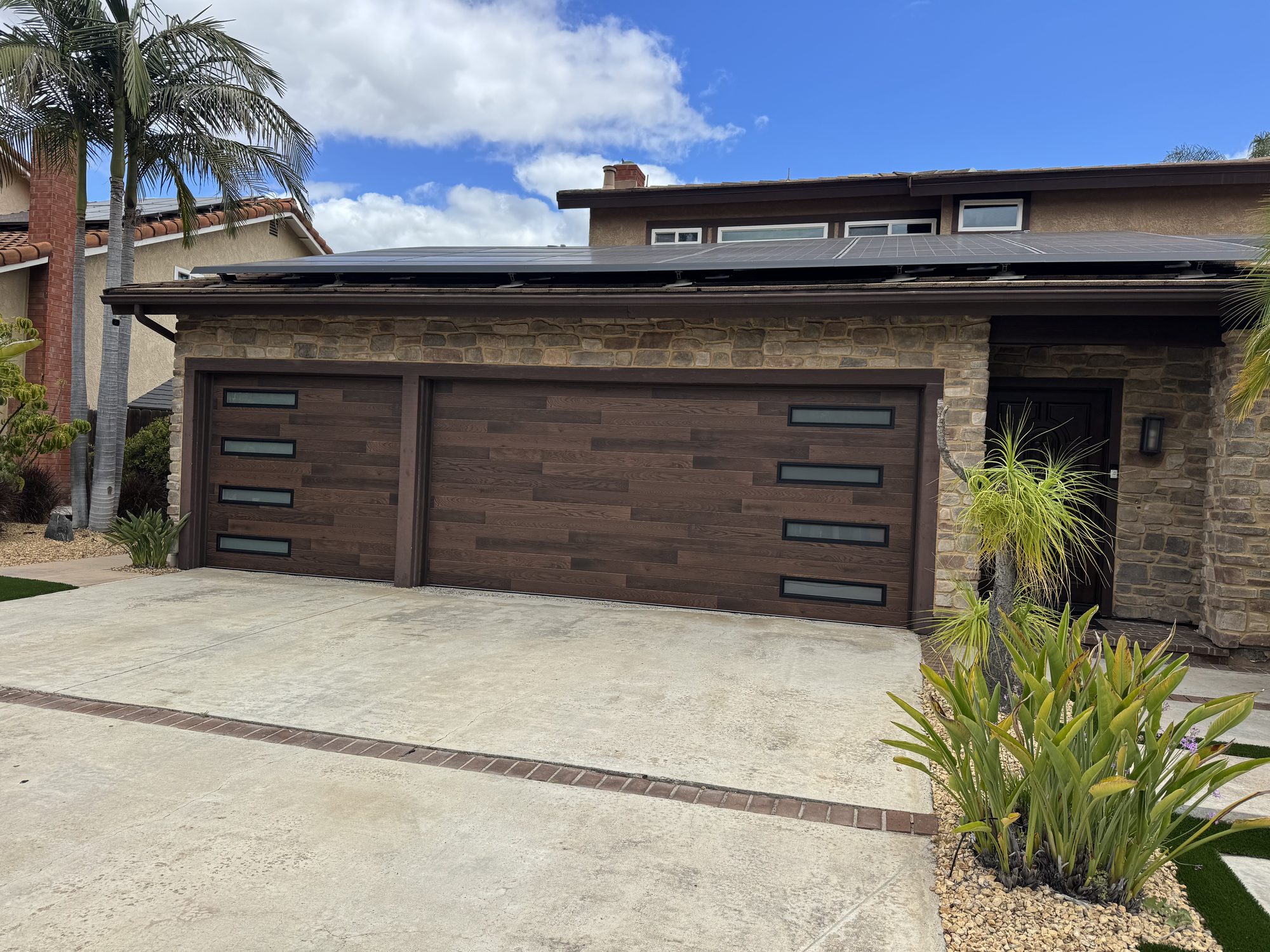 Clopay plank-style garage doors in a deep walnut finish with black-framed window slots, installed on a stone-front home in Poway, CA by A Reliable Garage Door